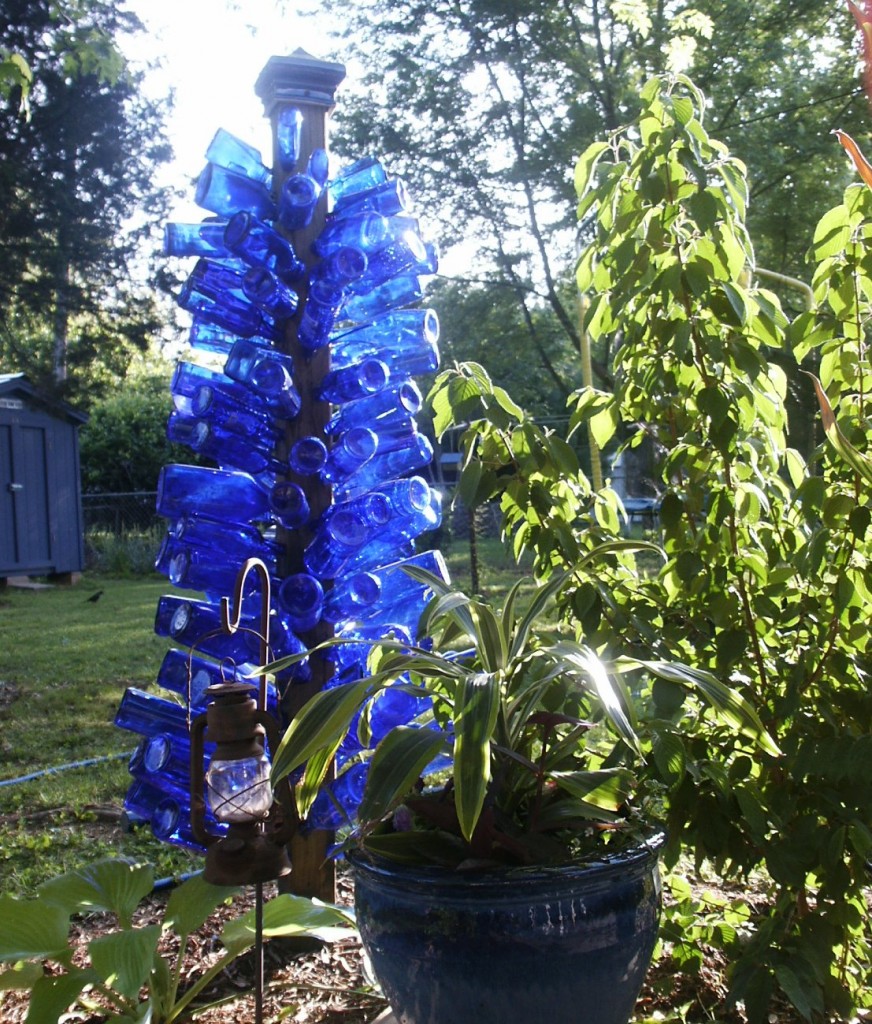 Picture of cobalt blue bottle tree with sun shining through the glass, next to shrubbery in a back yard garden.