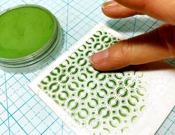 A finger applying green pastel to textured polymer clay.